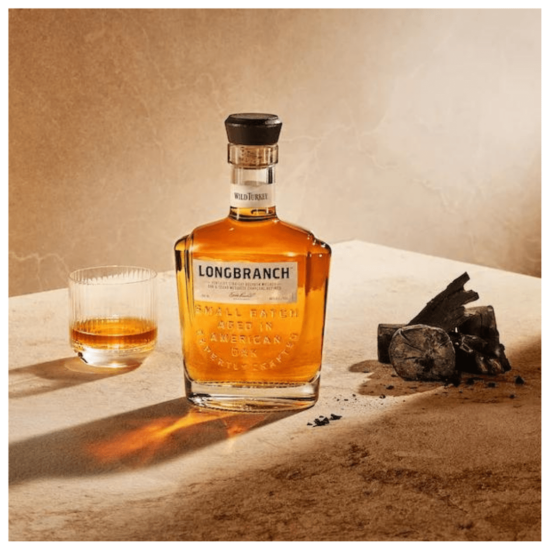 A photo of a bottle of Wild Turkey Longbranch bourbon and a glass of bourbon on a table. The bottle has a black label with gold text that says