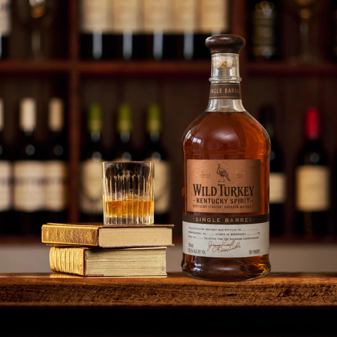 A bottle of Wild Turkey Single Barrel Kentucky Straight bourbon whiskey stands on a wooden bar counter next to a small glass of the amber liquid and a stack of two books, with a blurred liquor shelf in the background.